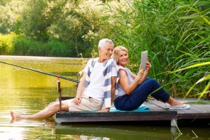 Portrait of elderly couple relaxing at nature. Senior woman and old man sitting back to back on the pier at lakeside while making self portrait with digital tablet.